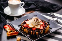 From above cappuccino in white mug on table with plate of round waffle with banana and strawberry topped with chocolate sauce and whipped cream — Stock Photo