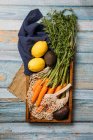 Vista dall'alto delle carote mature raccolte con fogliame verde, limone e avocado fresco poste sul tagliere sul tavolo di legno con shopping bag sostenibile — Foto stock