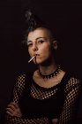 Доросла жінка з моховиком і пірсингом дивиться на камеру і запалює сигарету з запальничкою на чорному тлі — стокове фото