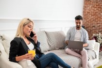 Mujer rubia positiva con taza de jugo hablando en el teléfono inteligente y sentado en el sofá cerca de novio étnico escribiendo en el teclado del ordenador portátil en la sala de estar del apartamento moderno - foto de stock