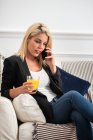 Mujer rubia feliz en ropa casual disfrutando de jugo de naranja fresco y el teléfono inteligente de navegación mientras está sentado en el sofá en casa - foto de stock