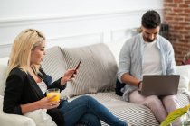 Femme blonde positive avec tasse de jus de navigation smartphone et assis sur le canapé près de copain ethnique tapant sur clavier d'ordinateur portable dans le salon de l'appartement moderne — Photo de stock