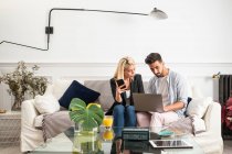 Positive blond woman with cup of juice browsing smartphone and sitting on couch near ethnic boyfriend typing on laptop keyboard in living room of modern apartment — Stock Photo