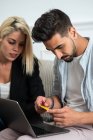 Jovem casal multiétnico sentado no sofá e fazendo compras on-line, juntamente com cartão de crédito em um laptop — Fotografia de Stock