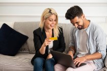 Задоволена блондинка посміхається і читає дані кредитної картки веселому етнічному хлопцю з ноутбуком, сидячи на дивані і роблячи онлайн-покупки разом — стокове фото