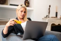 Mulher loira em roupas casuais entrando credenciais de cartão de crédito no laptop enquanto sentada em poltrona confortável e fazendo compras on-line em casa — Fotografia de Stock