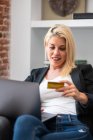 Блондинка-жінка в повсякденному одязі, що входить в облікові дані кредитних карток на ноутбуці, сидячи в зручному кріслі і роблячи онлайн-покупки вдома — стокове фото