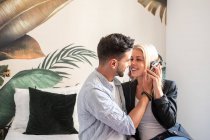 Веселая женщина в наушниках улыбается и пытается поцеловать этнического парня, слушая музыку дома вместе — стоковое фото