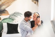 Весела жінка в навушниках посміхається і намагається поцілувати етнічного хлопця, слухаючи музику вдома разом — стокове фото