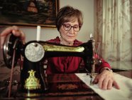 Senior lady in glasses using retro sewing machine to create linen napkin in cozy room at home — Stock Photo