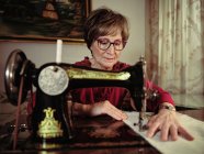 Senior senhora em óculos usando máquina de costura retro para criar guardanapo de linho no quarto acolhedor em casa — Fotografia de Stock