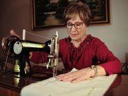 Seniorin in Brille fertigt mit Retro-Nähmaschine Leinen-Serviette in gemütlichem Zimmer zu Hause — Stockfoto