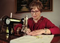 Senior lady in glasses using retro sewing machine to create linen napkin in cozy room at home — Stock Photo
