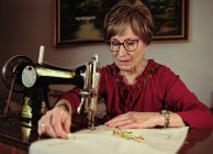 Señora mayor en gafas utilizando la máquina de coser retro para crear servilleta de lino en la habitación acogedora en casa - foto de stock