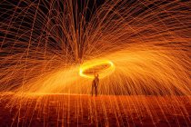 Silueta de larga exposición de persona irreconocible girando palo ardiente emitiendo destellos mientras baila con fuego por la noche - foto de stock