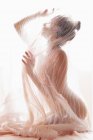 Vista lateral de modelo anónimo desnudo cubierto con tela plisada transparente de cortina contra la luz solar brillante - foto de stock