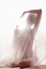 Anonymous naked model covered with transparent pleated fabric of curtain against bright sunlight — Stock Photo