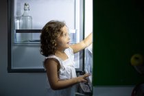 Маленька дівчинка в сплячому одязі шукає закуску всередині відкритого холодильника вночі на кухні вдома — стокове фото