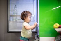 Side view of cute little child standing on stool and taking food from open refrigerator in cozy kitchen at home — Stock Photo