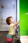 Vista laterale di bambino carino in piedi su sgabello e prendere il cibo dal frigorifero aperto in cucina accogliente a casa — Foto stock