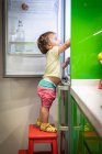 Side view of cute little child standing on stool and taking food from open refrigerator in cozy kitchen at home — Stock Photo