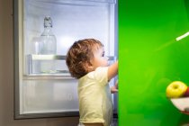 Seitenansicht des niedlichen kleinen Kindes, das auf einem Hocker steht und in der gemütlichen Küche zu Hause Lebensmittel aus dem offenen Kühlschrank nimmt — Stockfoto