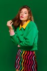 Young blonde woman model in stylish colorful outfit looking at camera thoughtfully while standing against green background — Stock Photo