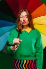 Young blonde female model in colorful outfit holding multicolored umbrella grimacing with closed eyes while standing against green background — Stock Photo