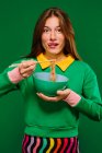 Positivo jovem fêmea no verde camisa olhando para câmera sorridente furar língua para fora enquanto comer gostoso instante macarrão com pauzinhos no verde fundo — Fotografia de Stock