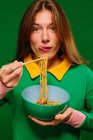 Positive young female in green shirt looking at camera grimacing sticking tongue out while eating yummy instant noodles with chopsticks on green background — Stock Photo