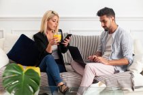 Femme blonde positive avec tasse de jus de navigation smartphone et assis sur le canapé près de copain ethnique tapant sur clavier d'ordinateur portable dans le salon de l'appartement moderne — Photo de stock