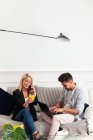 Positive blonde Frau mit einer Tasse Saft, die auf dem Smartphone spricht und neben ihrem Freund auf der Couch sitzt und auf der Laptop-Tastatur im Wohnzimmer einer modernen Wohnung tippt — Stockfoto
