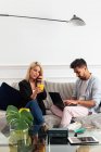 Позитивна блондинка з чашкою соку, що говорить на смартфоні і сидить на дивані біля етнічного хлопця, друкуючи на клавіатурі ноутбука у вітальні сучасної квартири — стокове фото