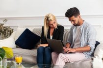 Blonde Frau und bärtiger Mann in lässiger Kleidung sitzen zu Hause auf dem Sofa und surfen gemeinsam am Laptop — Stockfoto