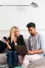 Positive blonde Frau surft im Smartphone und sitzt auf der Couch neben ihrem Freund und tippt auf der Laptop-Tastatur im Wohnzimmer einer modernen Wohnung — Stockfoto
