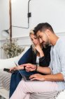 Positive blonde Frau surft im Smartphone und sitzt auf der Couch neben ihrem Freund und tippt auf der Laptop-Tastatur im Wohnzimmer einer modernen Wohnung — Stockfoto