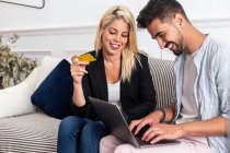 Delighted blonde female smiling and reading credit card credentials to cheerful ethnic boyfriend with laptop while sitting on sofa and making online purchases together — Stock Photo
