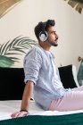 Side view of thoughtful bearded Hispanic male in headphones sitting on bed and enjoying good music at home — Stock Photo