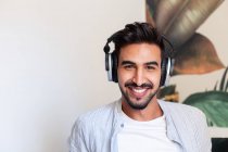 Щасливий етнічний хлопець в навушниках посміхається і дивиться на камеру, слухаючи музику вдома — стокове фото