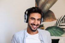 Glücklicher ethnischer Typ mit Kopfhörern, der lächelt und in die Kamera schaut, während er zu Hause Musik hört — Stockfoto