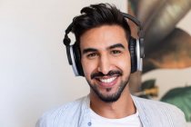 Happy ethnic guy in headphones smiling and looking at camera while listening to music at home — Stock Photo