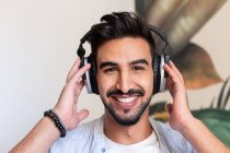 Glücklicher ethnischer Typ mit Kopfhörern, der lächelt und in die Kamera schaut, während er zu Hause Musik hört — Stockfoto