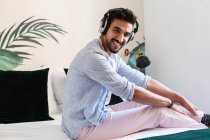 Seitenansicht eines nachdenklichen bärtigen hispanischen Mannes mit Kopfhörern, der auf dem Bett sitzt und zu Hause gute Musik genießt und in die Kamera schaut — Stockfoto