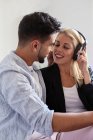 Весела жінка в навушниках посміхається і намагається поцілувати етнічного хлопця, слухаючи музику вдома разом — стокове фото