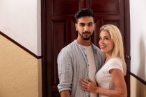 Positiv vielfältige Mann und Frau umarmen einander und blicken in die Kamera, während sie gegen flache Tür stehen — Stockfoto