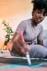 Зверху урожаю анонімні чорні жінки з пензлем малюють барвисті картини на папері, сидячи на підлозі вдома — стокове фото