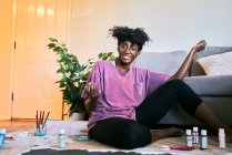 From above of crop anonymous black female with paintbrush drawing colorful picture on paper while sitting on floor at home — Stock Photo