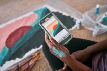 Зверху урожаю анонімні чорні жінки з пензлем малюють барвисті картини на папері, сидячи на підлозі вдома — стокове фото