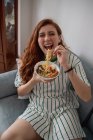 From above cheerful redhead female laughing and picking noodles from bowl of tasty ramen while sitting on couch at home — Stock Photo