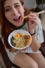 From above cheerful redhead female laughing blinking eye and picking noodles from bowl of tasty ramen while sitting on couch at home — Stock Photo